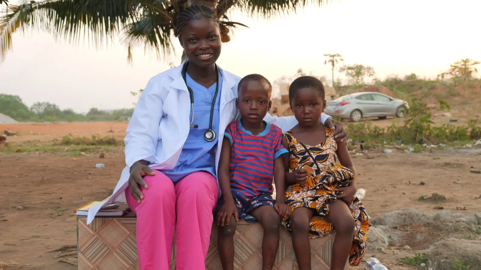 Smiling health worker in a white coat sitting outdoors with two young children